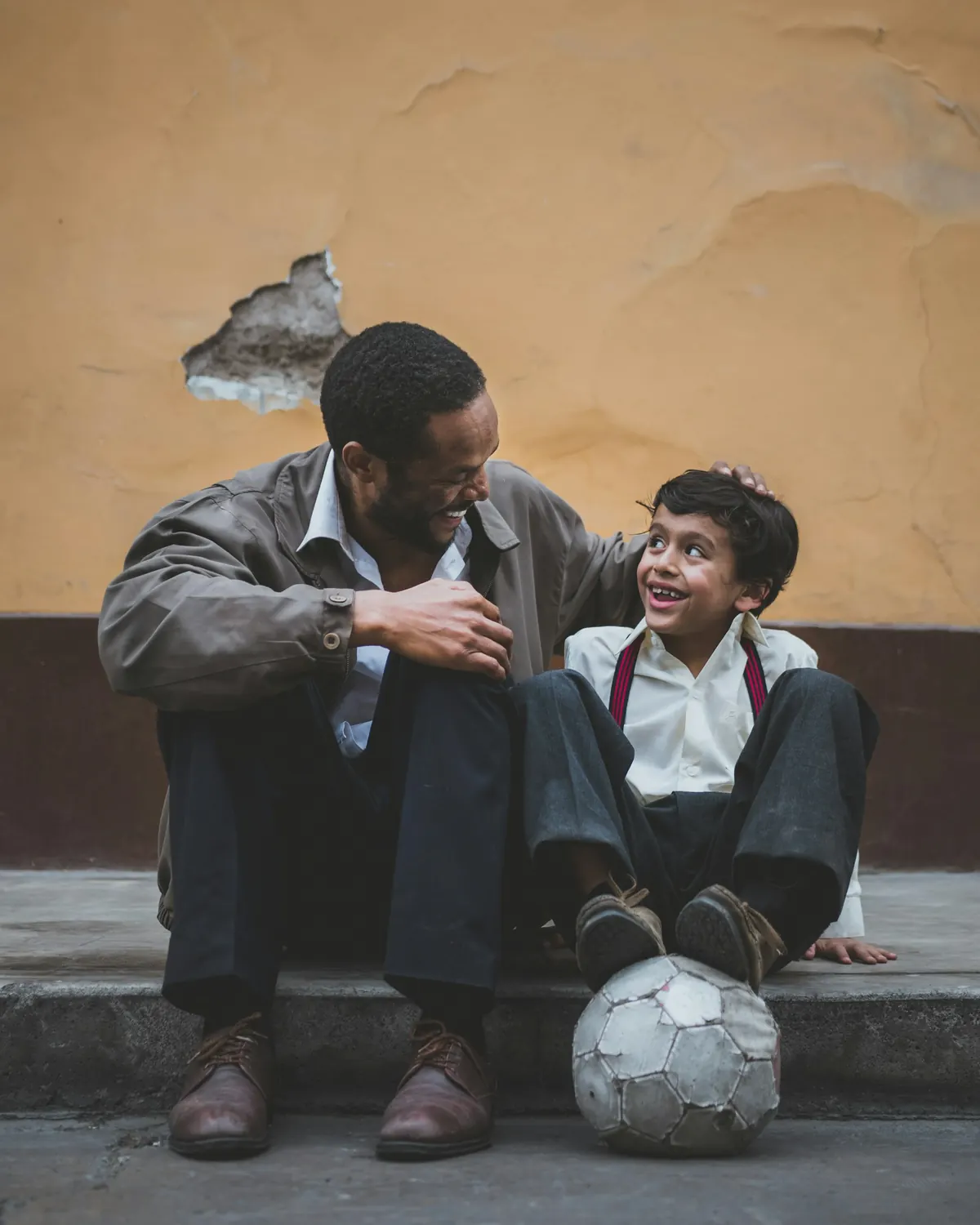 Un papá y su hijo platican sentados en una banqueta, con un balón de futbol